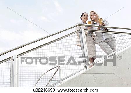 Low angle view of young businesswomen holding disposable coffee cups while standing by railing against sky View Large Photo Image Stock Photo - Low angle view of young businesswomen holding disposable coffee cups while standing by railing against sky. Fotosearch
