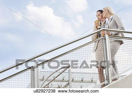 Low angle view of young businesswomen with disposable coffee cups looking away while standing by railing against sky View Large Photo Image Stock Photo - Low angle view of young businesswomen with disposable coffee cups looking away while standing by railing against sky. Fotosearch