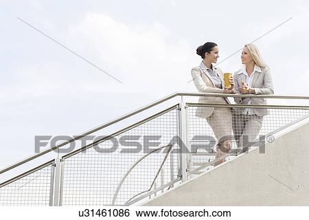Stock Photograph - Low angle view of young businesswomen talking while standing by railing against sky. Fotosearch