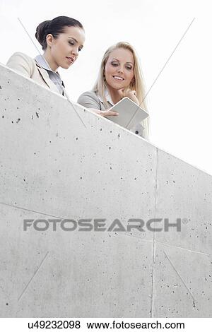 Stock Photo - Low angle view of young female business executives using digital tablet while standing on terrace against sky. Fotosearch