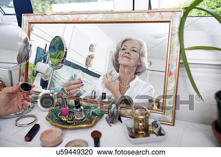 Stock Image - Mirror reflection of senior woman spraying perfume on herself at home. Fotosearch