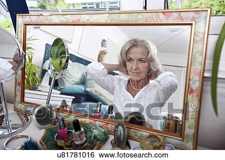 Stock Photograph - Mirror reflection of senior woman putting on necklace at home. Fotosearch