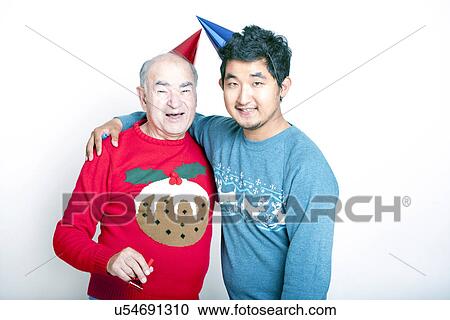 Stock Image - Portrait of a Senior adult man and a young Asian man wearing Christmas jumpers and party hats. Fotosearch