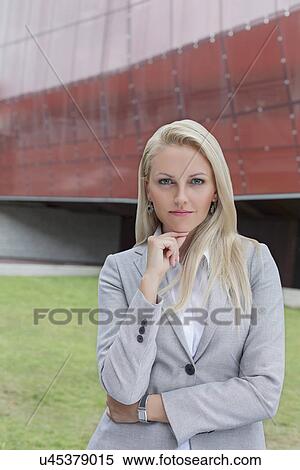 Stock Photography - Portrait of confident businesswoman standing with hand on chin against office building. Fotosearch