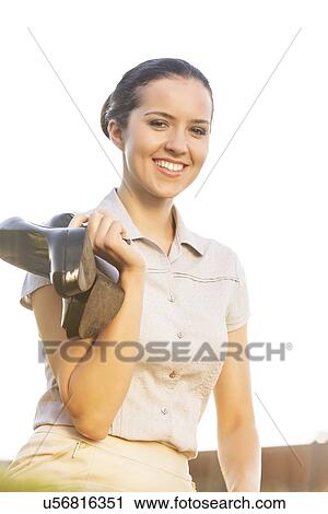 Stock Image - Portrait of happy young businesswoman holding high heels against sky. Fotosearch