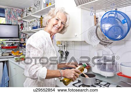 Picture - Portrait of senior woman pouring olive oil to saucepan in domestic kitchen. Fotosearch