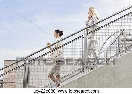 Profile shot of businesswomen holding disposable cup and mobile phone while moving down stairs together against sky View Large Photo Image Stock Photo - Profile shot of businesswomen holding disposable cup and mobile phone while moving down stairs together against sky. Fotosearch