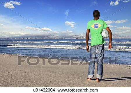Picture - Rear view of man standing wearing Brazilian t shirt with football on a beach. Fotosearch