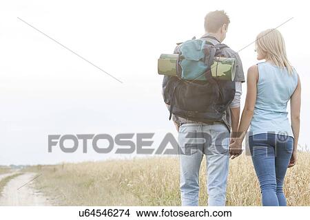 Picture - Rear view of young hiking couple holding hands while walking in countryside. Fotosearch