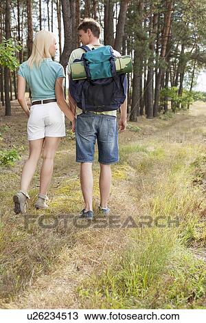 Stock Image - Rear view of young hiking couple holding hands while walking in forest. Fotosearch