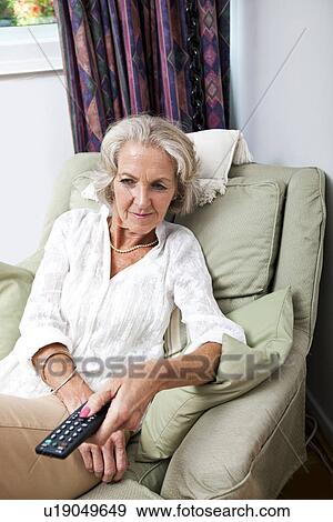 Stock Photo - Senior woman changing channels with remote control on armchair at home. Fotosearch