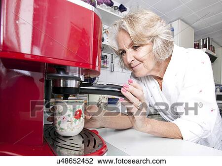Stock Image - Senior woman dispensing coffee from machine at kitchen counter. Fotosearch