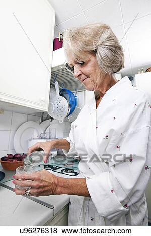 Stock Photo - Senior woman pouring water in glass while standing at kitchen counter. Fotosearch