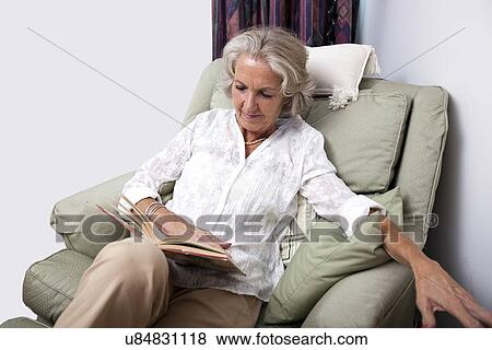 Stock Photo - Senior woman reading book while relaxing on armchair at home. Fotosearch