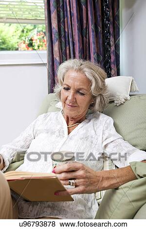 Stock Photo - Senior woman with coffee cup reading book while relaxing on armchair at home. Fotosearch