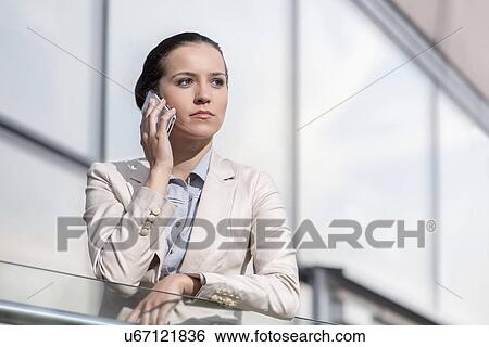 Serious young businesswoman using cell phone at office railing View Large Photo Image Stock Photograph - Serious young businesswoman using cell phone at office railing. Fotosearch