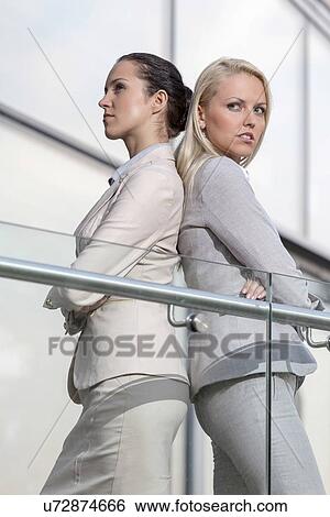 Serious young businesswomen standing back to back at office railing View Large Photo Image Stock Photograph - Serious young businesswomen standing back to back at office railing. Fotosearch