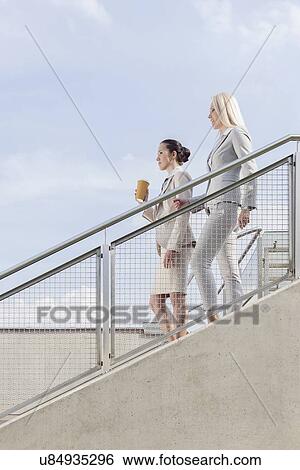 Stock Photograph - Side view of businesswomen moving down stairs against sky. Fotosearch