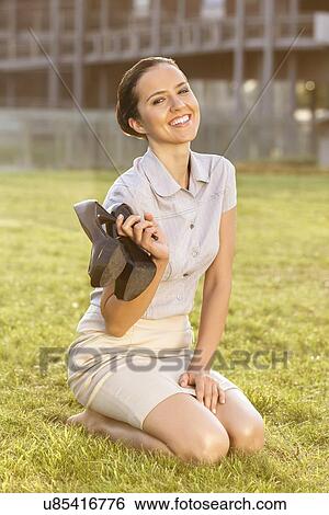 Stock Photograph - Smiling young businesswoman holding high heels while sitting in office lawn. Fotosearch