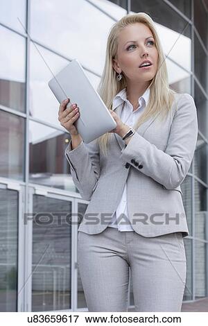Stock Photo - Thoughtful businesswoman holding digital tablet while looking away against office building. Fotosearch