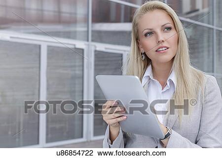 Stock Image - Thoughtful young businesswoman using digital tablet while looking away against office building. Fotosearch