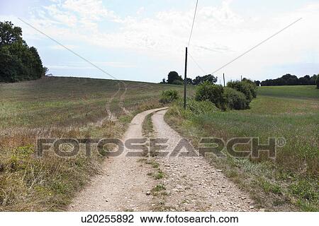 Stock Image - Unpaved road in grassy field against cloudy sky. Fotosearch