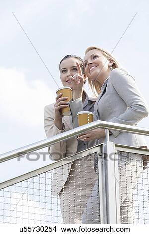 Young businesswoman with disposable coffee cup pointing while standing with coworker by railing against sky View Large Photo Image Picture - Young businesswoman with disposable coffee cup pointing while standing with coworker by railing against sky. Fotosearch
