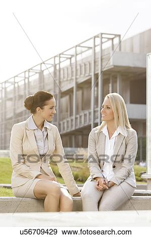 Young businesswomen looking at each other while sitting against office building View Large Photo Image Stock Photo - Young businesswomen looking at each other while sitting against office building. Fotosearch