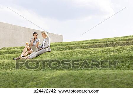 Young businesswomen using laptop together while sitting on grass steps against sky View Large Photo Image Stock Image - Young businesswomen using laptop together while sitting on grass steps against sky. Fotosearch