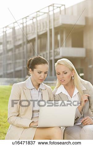 Stock Photo - Young businesswomen using laptop together while sitting against office building. Fotosearch