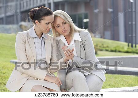 Young businesswomen using mobile phone together while sitting against office building View Large Photo Image Stock Image - Young businesswomen using mobile phone together while sitting against office building. Fotosearch
