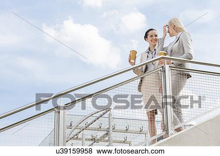 Young businesswomen with disposable coffee cups standing by railing against sky View Large Photo Image Stock Photo - Young businesswomen with disposable coffee cups standing by railing against sky. Fotosearch