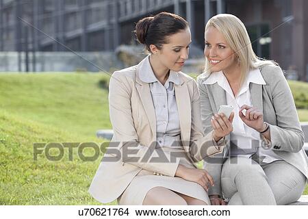 Picture - Young businesswomen with mobile phone sitting against office building. Fotosearch