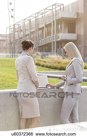 Stock Photograph - Young businesswomen working together on laptop while standing against office building. Fotosearch