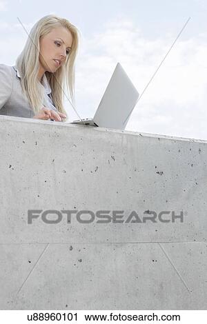 Stock Image - Young female business executive using laptop on terrace against sky. Fotosearch