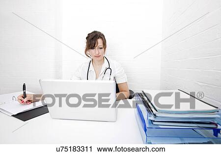 Young female doctor with laptop writing notes on desk in clinic View Large Photo Image Stock Image - Young female doctor with laptop writing notes on desk in clinic. Fotosearch