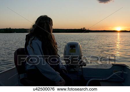 Stock Photograph - Girl sitting on a motorboat in the lake at sunset, Lake of The Woods, Keewatin, Ontario, Canada. Fotosearch