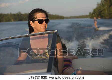Stock Image - Woman driving a motorboat with a person waterskiing, Lake of The Woods, Keewatin, Ontario, Canada. Fotosearch