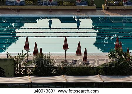 Stock Photo - Aerial view of athlete's swimming pool with channels marked by red and white dividers, with closed umbrellas silhouetted on red tiles alongside, trees reflected in water, early morning sunshine, Umbria, Italy. Fotosearch