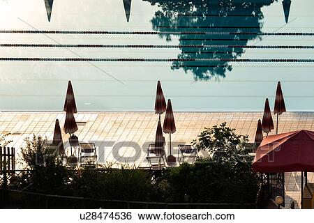 Stock Photograph - Aerial view of athlete's swimming pool with channels marked by red and white dividers, with closed umbrellas silhouetted on red tiles alongside, trees reflected in water, early morning sunshine, Umbria, Italy. Fotosearch