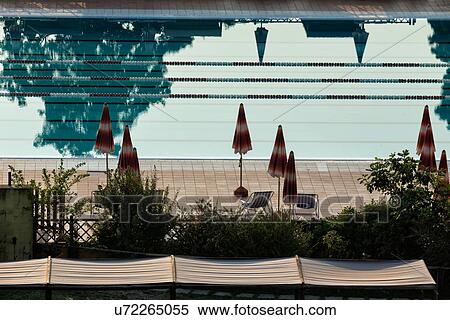 Stock Photography - Aerial view of athlete's swimming pool with channels marked by red and white dividers, with closed umbrellas silhouetted on red tiles alongside, trees reflected in water, early morning sunshine, Umbria, Italy. Fotosearch