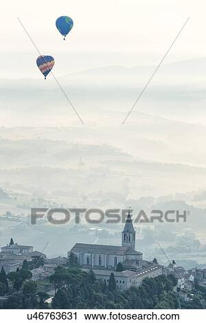 Stock Image - Aerial view of church and spire of San Fortunato church with cloister complex surrounding, Piazza del Popolo to the left, in medieval city of Todi, and layered hills of the Middle Tiber Valley in early morning hazy sunshine, with two hot air balloons floa. Fotosearch