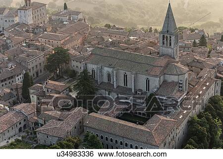 Stock Photograph - Aerial view of church and spire of San Fortunato church with cloister complex surrounding, Piazza del Popolo beyond, in medieval city of Todi in early morning hazy sunshine, Todi, Umbria, Italy. Fotosearch