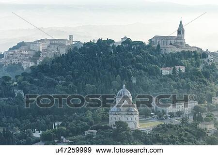 Stock Photo - Aerial view of medieval city of Todi with church and spire of San Fortunato church on right, and Duomo and Piazza del Popolo on left, Santa maria della Consolazione Tempio in the foreground, in early morning hazy sunshine, Todi, Umbria, Italy. Fotosearch