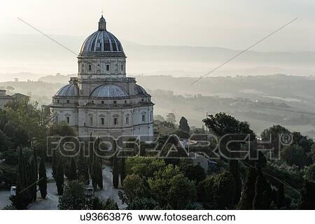 Stock Photograph - Aerial view of Santa Maria della Consolazione Tempio Church outside Todi, with surrounding trees and buildings with early morning hazy sunlight, layering the hills of the Middle Tiber Valley beyond, Umbria, Italy. Fotosearch