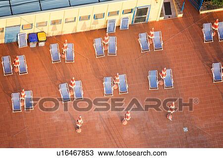 Aerial view of swimming pool blue deck chairs in pairs, on red tiles with closed red umbrellas, creating geometric pattern in close up, early morning sunshine, Umbria, Italy View Large Photo Image Stock Image - Aerial view of swimming pool blue deck chairs in pairs, on red tiles with closed red umbrellas, creating geometric pattern in close up, early morning sunshine, Umbria, Italy. Fotosearch