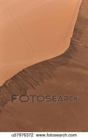 Stock Image - Close up view of line of red sand dune with different tones in shadow of early morning sunlight, and sand being blown by winds, Namib desert, Namibia, South West Africa. Fotosearch