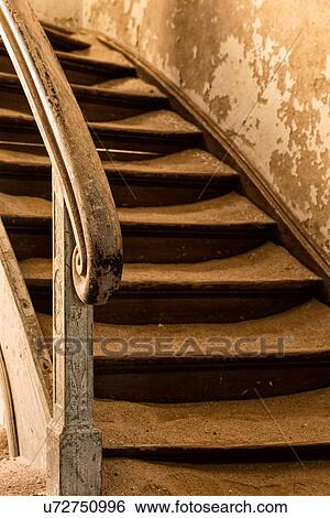 Stock Photograph - Kolmanskop interior view of hallway: curved wooden stairway with turned wooden balustrade, san over steps, abandoned German mining town, Namib desert, Namibia, South West Africa. Fotosearch