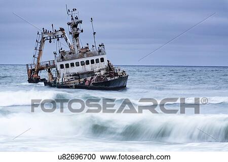Stock Image - Old fishing boat run aground in shallow water off Skeleton Coast: twilight with motion blur of waves splashing, Namibia, South West Africa. Fotosearch