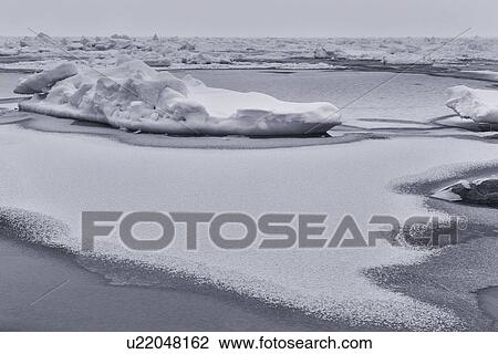 Stock Image - Rocks covered with snow at water's edge, ice patches forming in the frozen sea creating patterns and textures, monochrome image, Shiretoko Peninsula, Hokkaido, Japan. Fotosearch